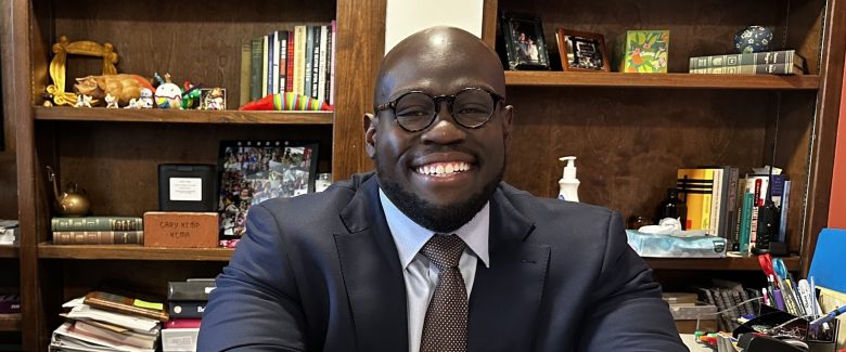 A black man in a suit sits at a desk with his hands crossed.