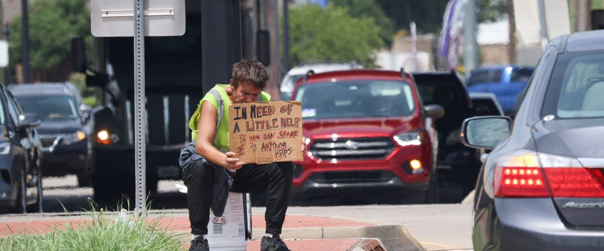 A man in a reflective vest sits on a white bucket and holds a sign.